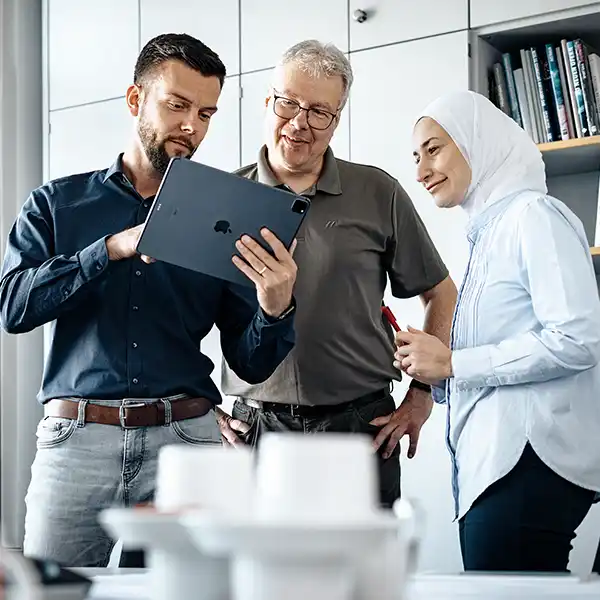 Drei Mitarbeitende - ein junger Mann, ein älterer Herr und eine Frau mit Kopftuch - schauen gemeinsam auf ein Tablet in einem modernen Büro